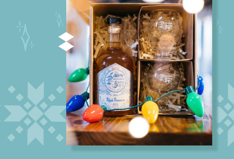 A box containing paper sawdust with a bottle of liquor and two short round glasses surround by paper packing material sit on a dark wood table. Surrounding the box is Christmas lights of various colors.