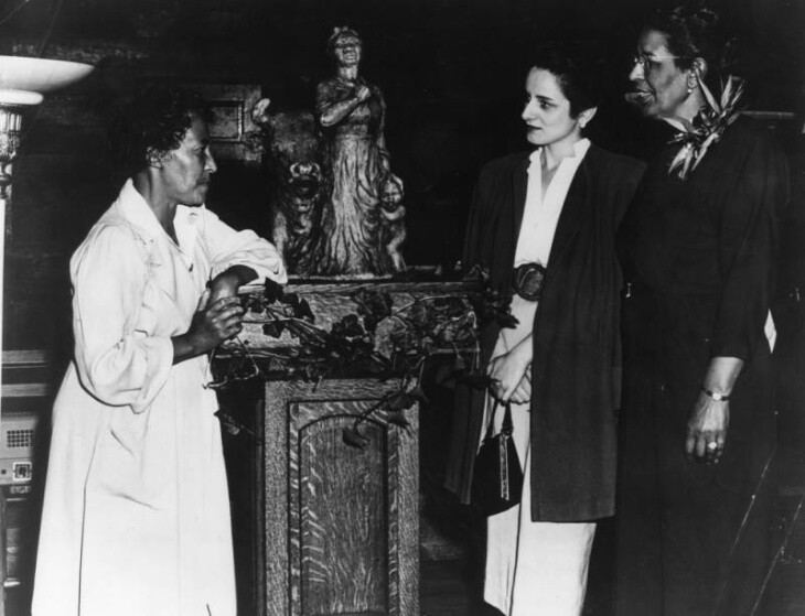 Three women gather around a statue on a wood stand.