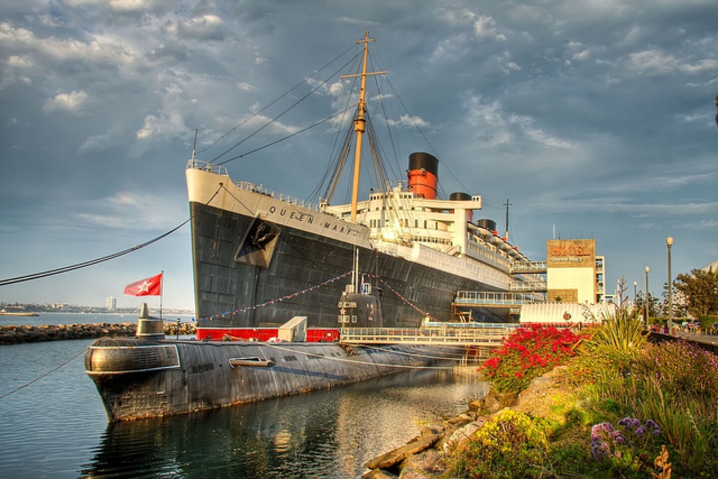 A ground-level view of the Queen Mary, docked in Long Beach, looking from the bow toward the back of the ship. The distinctive orange and black funnels are visible. A former Soviet submarine called the Scorpion is visible in the foreground, beside the ocean liner.