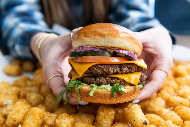 A multi-layer vegan burger with "cheese" tomato, onion and pickle is held by a person above tater tots