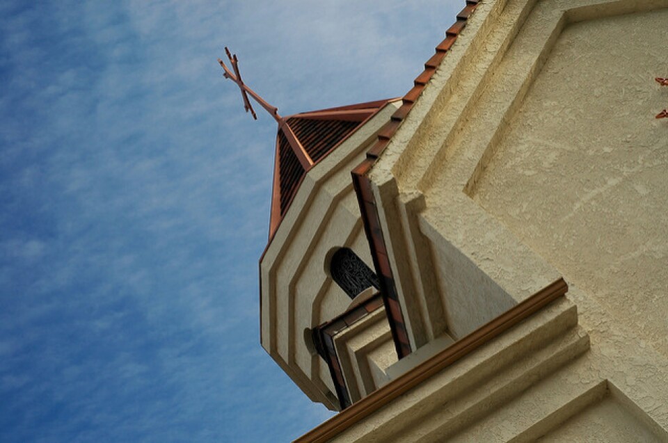 A cross sits at the top of a copper lined octagonal church dome against a cloud streaked blue sky.