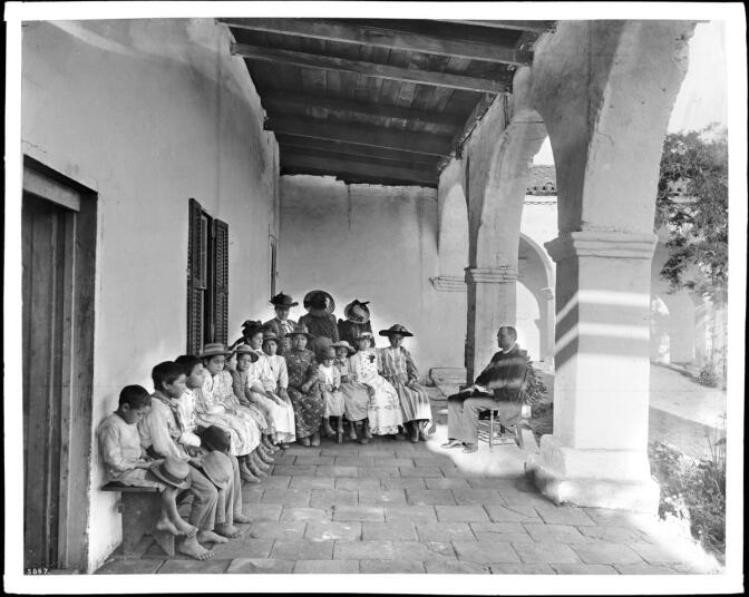 About 10 children and six women sit or stand in a quarter circle around the priest under the arcade. The priest, dressed in priest garb, sits in a wooden chair with a book held open in one hand. 