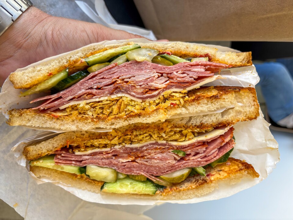 A hand with light skin tone holds a stacked sandwich cut in half. The sandwich is filled with thinly sliced deli meat , pickles and cucumbers,  a layer of crushed chips for added crunch, and cheese. It's made with thick slices of toasted bread and has a visible sauce