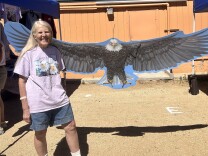 A woman with long hair and in a t-shirt with two eagles on it is standing next to a cut out of an eagle with spreading wings.