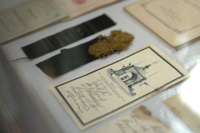 A close up of the funeral mementos. There is a strip of black cloth with dried brown moss on it. To the right is a card that describes the relic with a black and white illustration of an archway.