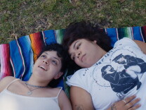 Two brown-skinned women lie in the grass on a colorful blanket. One woman looks at the other, while the other looks up to the sky. They both wear white T-shirts — one of their shirts has a black and white image of two women sitting with their legs entangled with one another.