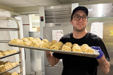 A man with medium skin tone — wearing a black t-shirt, a hat, glasses and purple latex gloves — carries a tray full of baked goods in an industrial kitchen.