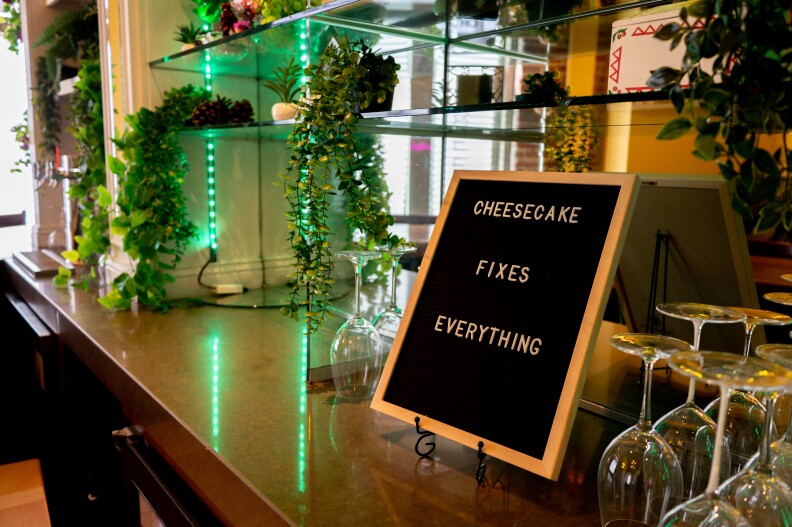 A sign on the counter reads "CHEESECAKE FIXERS EVERYTHING." It is surrounded by shelves, small plants, and empty upside-down wine glasses.