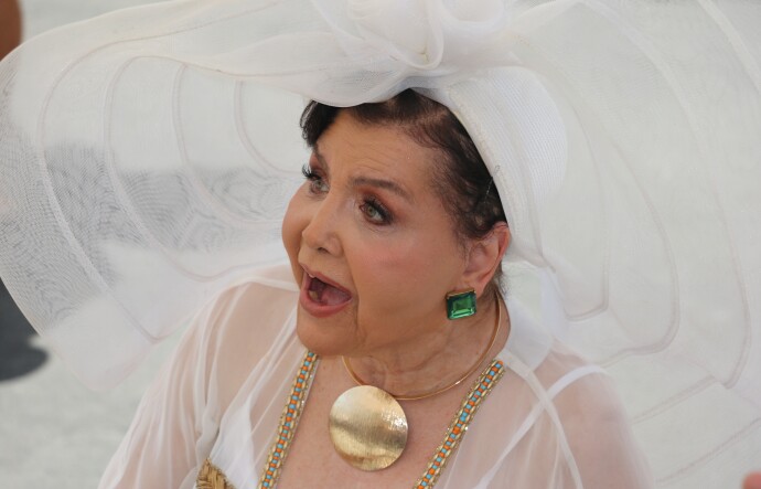 An older woman of mixed heritage wearing a large gold amulet necklace, big green gem earrings and an enormous-brimmed white hat.