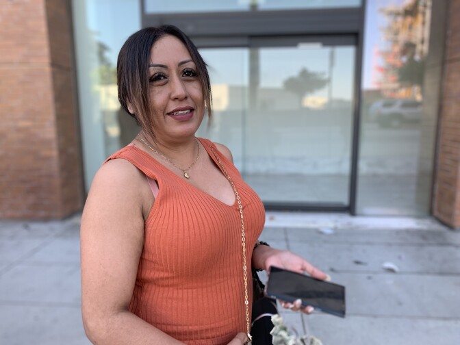 A portrait of a woman from the waist up who's standing outside. She's smiling at the camera with her phone in one hand.