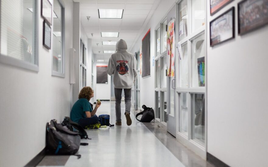 One student with light skin and shoulder length dark blonde hair sits next to a row of backpacks lined up in a school hallway holding a sandwich in front of his mouth as he looks up at a student walking past wearing dark gray pants and a light pullover sweatshirt with a red and purple circular logo over it. The student walking past has his back turned.