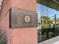 The exterior of a building has a terra cotta-colored stone wall with the words : County of Orange County Administration South on a plaque dated 2029. The names of county officials are on the sign. A glass wall reflects U.S. California and the county flag.