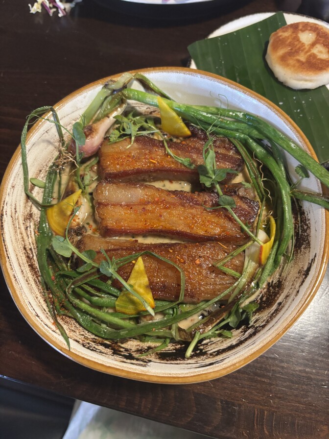 Round plate showing sliced lamb belly arranged in fan shape over green beans and greens, with lemon wedge and yellow squash blossoms as garnish