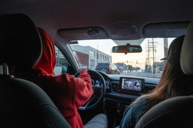 A person wearing a red hooded sweatshirt driving a car with a woman wearing a black baseball cap sitting next to them in the passenger seat. The two are seen from the vantage point of the backseat of a car.