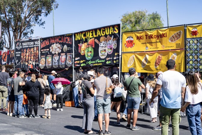 A crowd of people stand in front of a row of food vendor stands, ranging from takoyaki to mini pancakes.