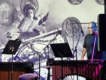 Ariel Campos plays a marimbas with a bass bow. He sports a black t-shirt and glasses. There is a multimedia art projection behind him. 