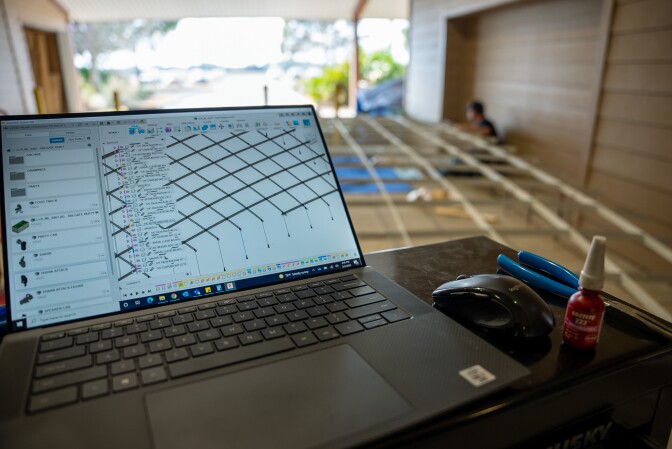 A laptop computer screen shows a criss crossing pattern on the right, various Legos on the left. Net to the computer on a workstation are a mouse, a vial, and pliers. The large roof of the mini stadium is being constructed in the background.