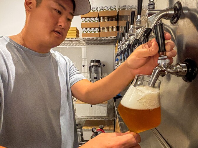 A man with medium dark skin tone in a light blue shirt and cap pours a golden beer from a tap into a tilted glass, creating a frothy head, with shelves of cans and equipment visible in the background.