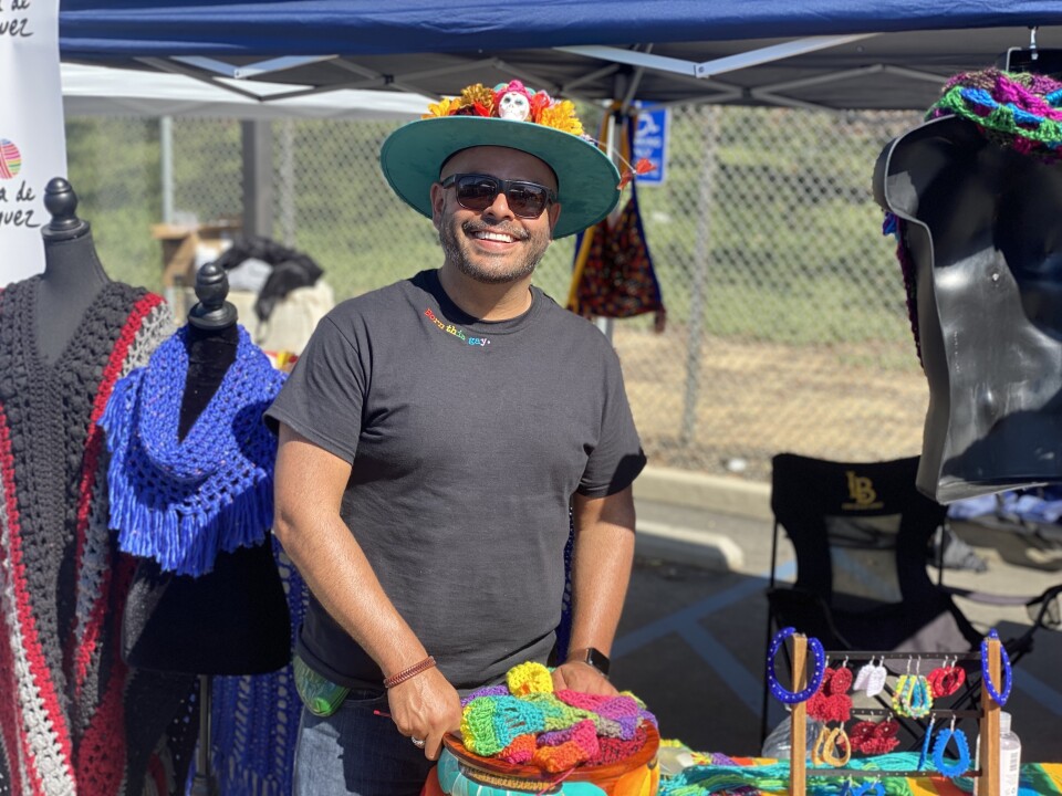 A man with a dark shirt and a hat with calabera decorations at a booth with knitting items at Queer Mercado.