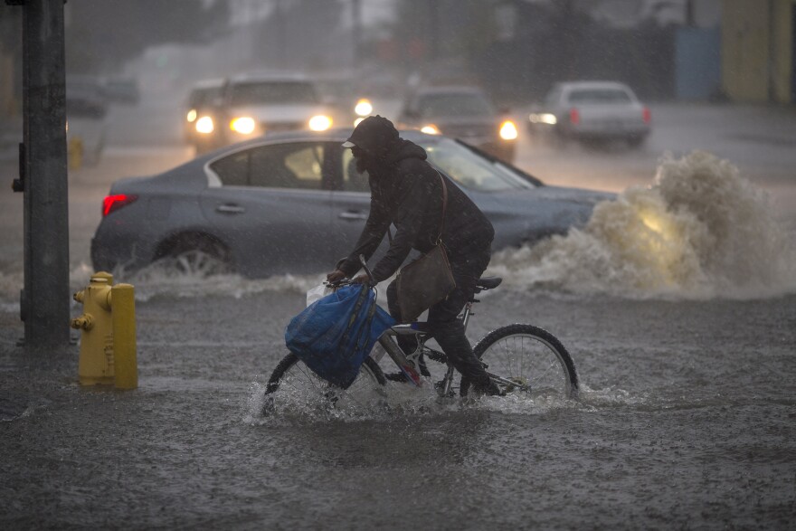 A cyclist rides through floodwater in the foreground as a car does the same in the background.