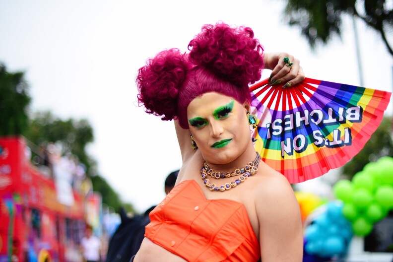 A fair-skinned person poses in the middle of a pride parade. They're wearing an orange crop top, green lipstick and eye makeup with magenta-colored hair. A rainbow fan is spread open in their hand.