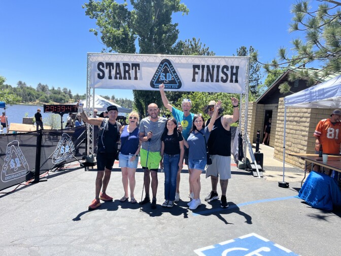 A group of people stand at a finish line with their arms around each other and up in the air. They are smiling.