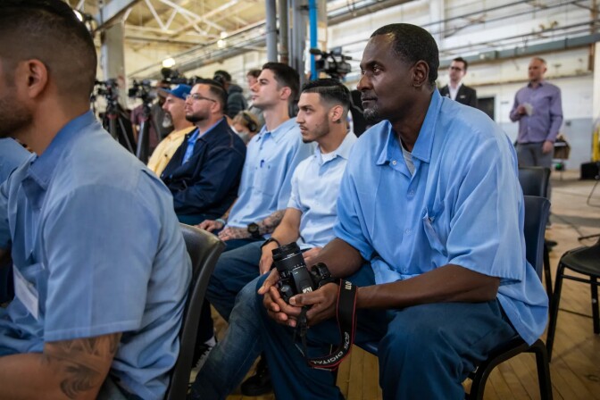 Several rows of men are seated inside what appears to be a warehouse. They are dressed mostly in blue collared short-sleeved shirts. The man closest to the front of the frame is Black and appears to be in his 40s or 50s. Rows of cameras are blurred in the background behind the men. 