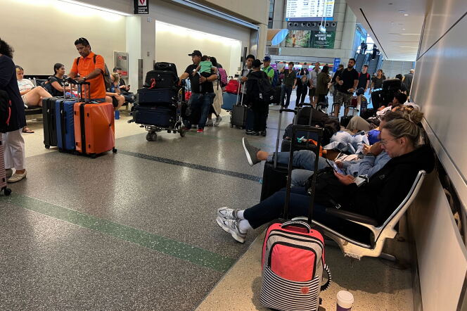 People push luggage carts and roller suitcases while others sit on chairs looking at their phone with their luggage beside them. A man wearing a dark t-shirt carries a child wearing a green t-shirt. 