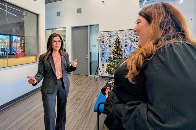 Two women chatting. Woman on left is wearing a grey suit and peach blouse, standing facing a woman wearing a black blouse.