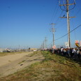 A long line of protester carrying signs that read "ICE OUT!" stand under power lines opposite a detention center on the other side of a concrete drainage channel.