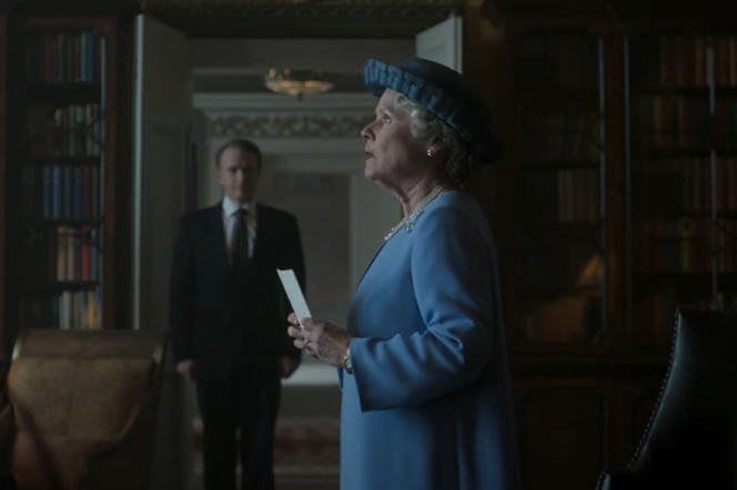 A profile of woman in a blue dress and blue hat holding a piece of paper as a man in a black suit in the distance looks at her. Book shelves line the walls.