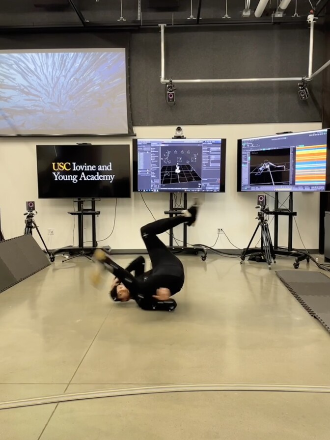 A wide view of a man with a medium-light skin tone as he spins on the ground dancing indoors. He's wearing a full blacksuit in front of cameras and large TV screens showing his body movement.