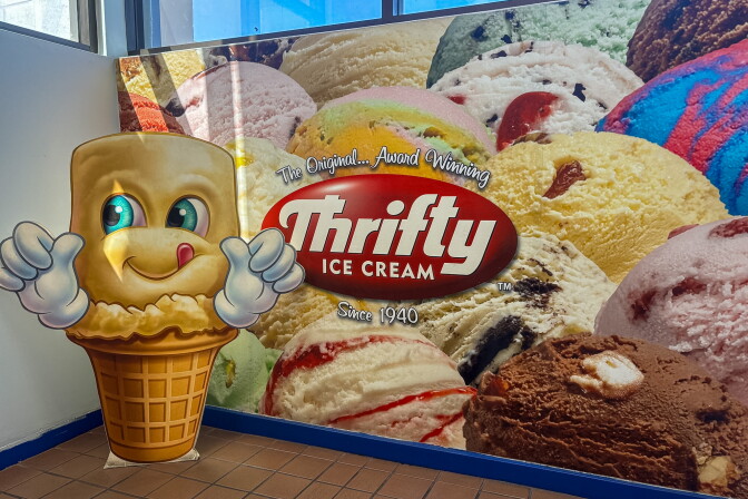 A life-sized cardboard cutout of Thrifty Ice Cream's smiling cone mascot giving two thumbs up, standing in front of a wall mural of colorful ice cream scoops and the Thrifty Ice Cream logo, "Since 1940."