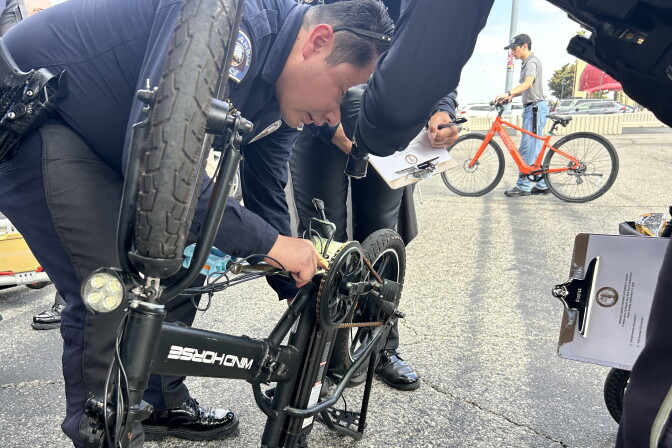 Police officers examine a folded electric bike's gears in a parking lot as another writes on a clipboard.