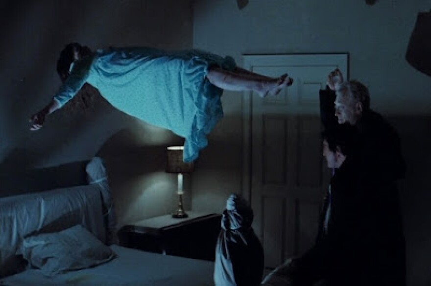 A young girl, Linda Blair, floats above a bed in a small bedroom. Blair wears a white night gown. Her arms are outstretched , her legs slightly together as she looks like she's laying down but in the air. Two men in all black stand at the foot of the bed, Max von Sydow and Jason Miller. von Sydow has one arm raised above his head with something in it. The room is dark, the only light coming from a lamp on the nightstand next to the bed. The room has a bluish glow to everything. 