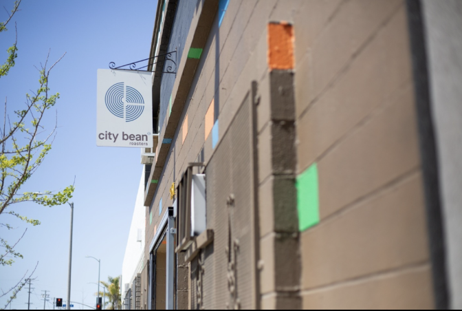 A brushed concrete façade dotted with pops of color sets the tone outside City Bean Roasters, where a clean white sign swings gently in the breeze.