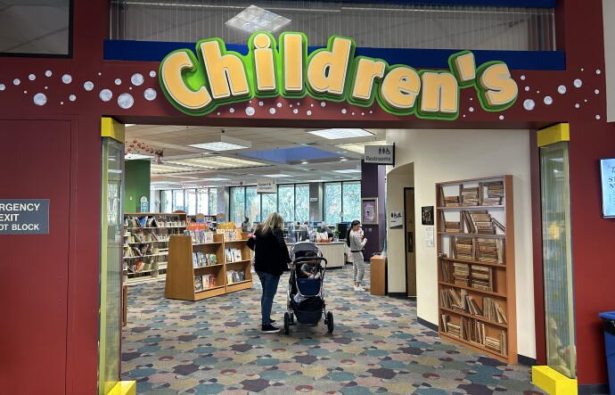 An entryway with a large yellow sign above that reads "Children's." Through the entry, there's a female-presenting person next to a baby stroller and shelves of books in the distance. 