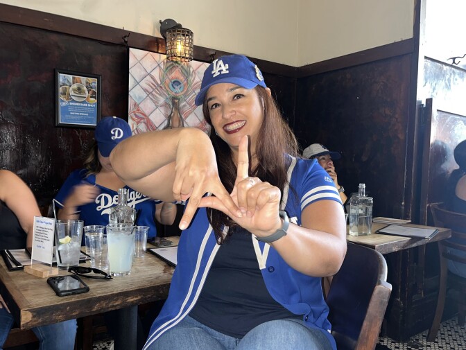 Latina fan wearing Dodgers jersey makes Dodgers symbol with fingers inside bar.