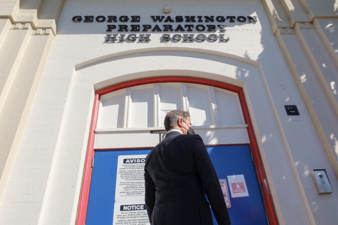 A man in a suit stands in front of a set of blue double doors, outside a school. He has his back turned to the camera.