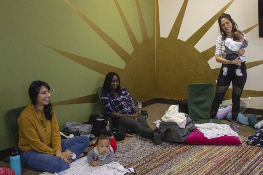 A woman in a mustard yellow long-sleeved shirt sits with her infant son. To her right is a pregnant woman in a blue plaid shirt and standing to her right is a woman with black leggings on holding an infant. The wall behind them is painted olive and cream with a large gold sun.