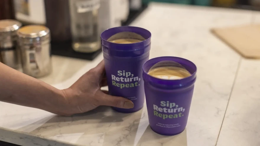 A hand entering the frame from the right holds a purple cup of coffee with foamed milk as it rests beside a shorter version of itself on a white marble table.