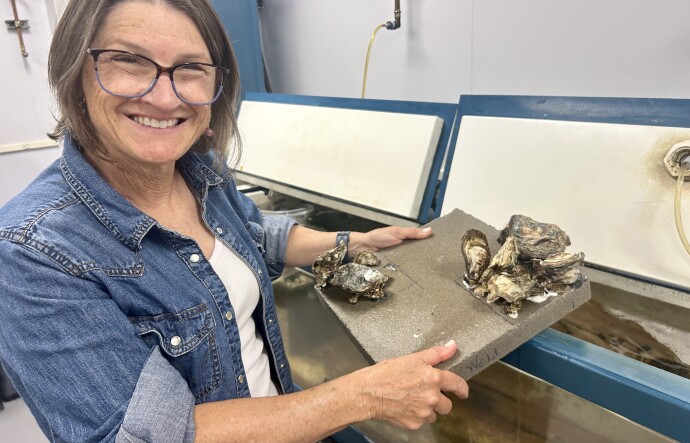 A woman with short brown hair, glasses and a jean jacket smiles into the camera while holding a small piece of concrete with oyster shells attached to it. She's standing in a room with several aquarium tanks.
