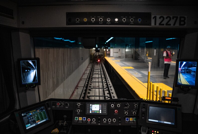 The cockpit of a subway train with a view of the track and the yellow-edged station platform as it arrives at a station. The instrument panel sits below the main viewport, and on the left and right sides of that viewport are vertical video screens. Above the viewport is a series of lights or dials and a placard that reads "1227B."