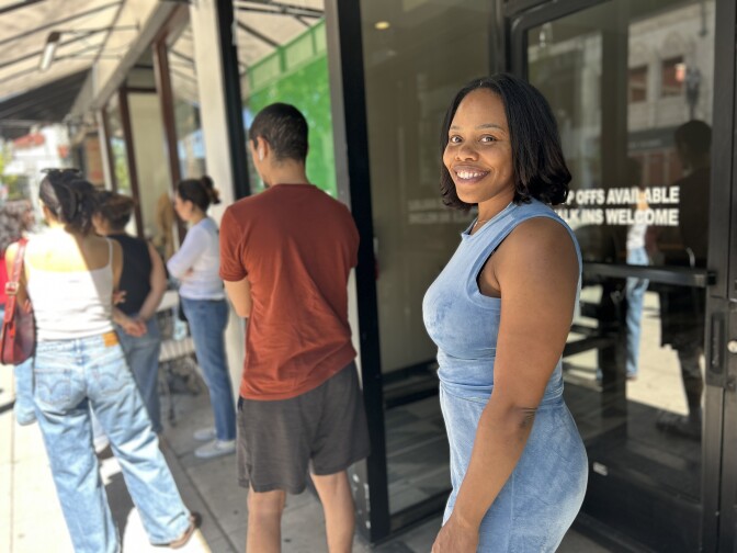 A young to middle aged woman with dark brown skin and dark brown short straight hair smiles under an awning. She is in a line of people. 