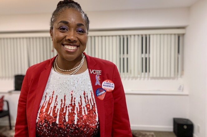 A woman with dark skin tone and hair tied back in a bun smiles and wears a red blazer with a red and white sequin shirt underneath. There are three pins on her left side of her jacket. The word Vote in rhinestones, a Kamala Harris for President 2024 button and red, white, and blue ribbon. 