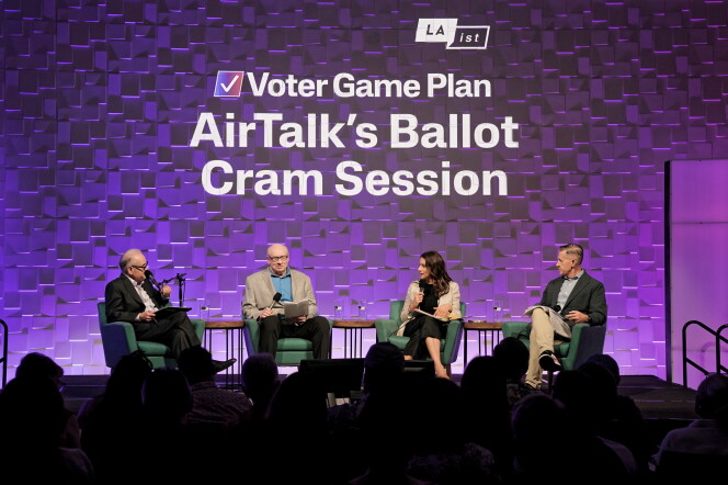 A photograph of a crowded, darkend room: The stage is dramatically lit in purple and there are four people sitting on stage in green club chairs, holding microphones.