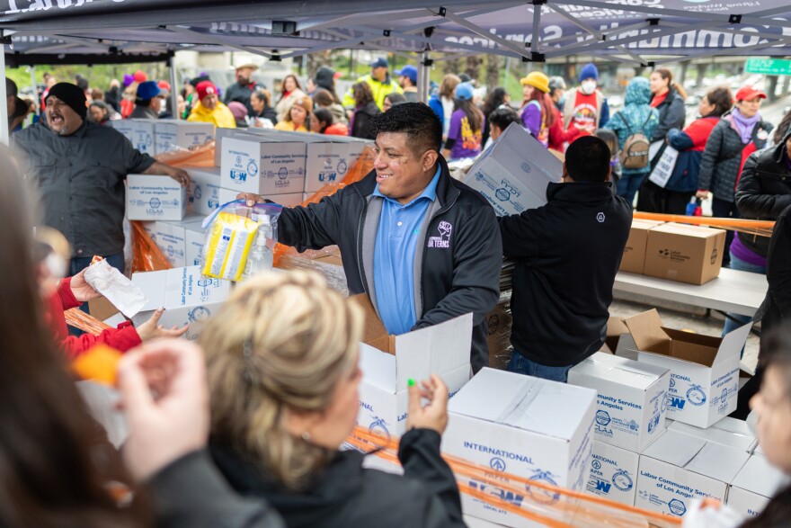 A man with light brown skin hands out a plastic bag with snacks in it. He is surrounded by boxes of food and stands under a tent. There's a large crowd nearby.