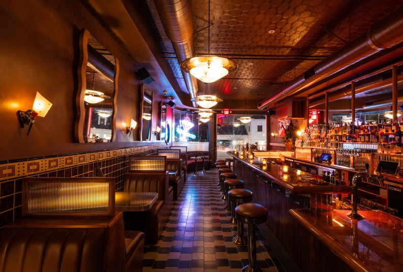 Interior of a warmly-lit restaurant featuring a bar with candles lit on the bar top and small booths on the opposite side of the room.