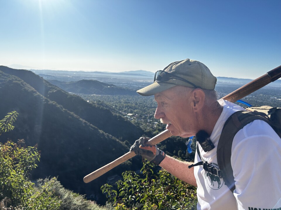 An older white man from the side carries a wooden handled tool with gloves, wears a white t-shirt and beige baseball cap. In the background is a vista of mountains and green and city in the distance. 