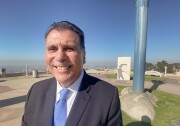 A man in a dark suit and blue tie smiles for the camera. Behind him we see a park and some sort of public monument.